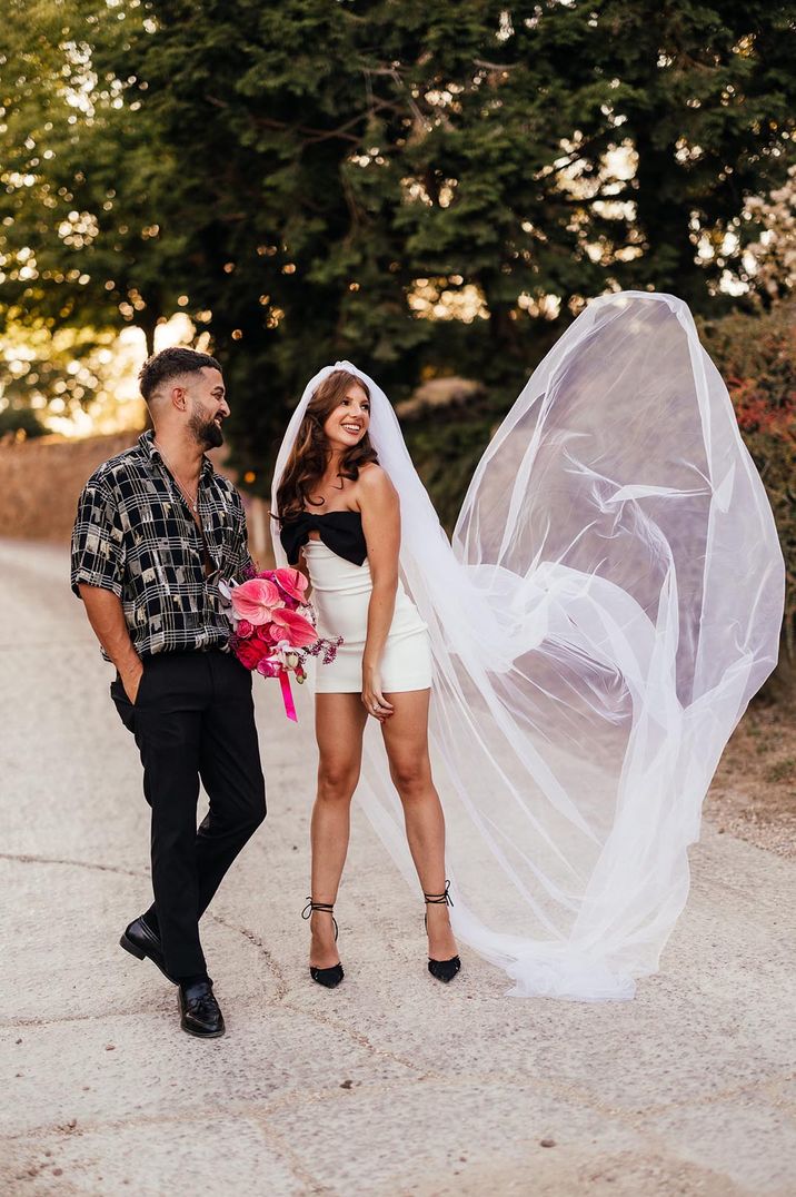 The groom in a patterned shirt and trousers stands with the bride in a short black and white bow wedding dress with cathedral length veil and pink bouquet 