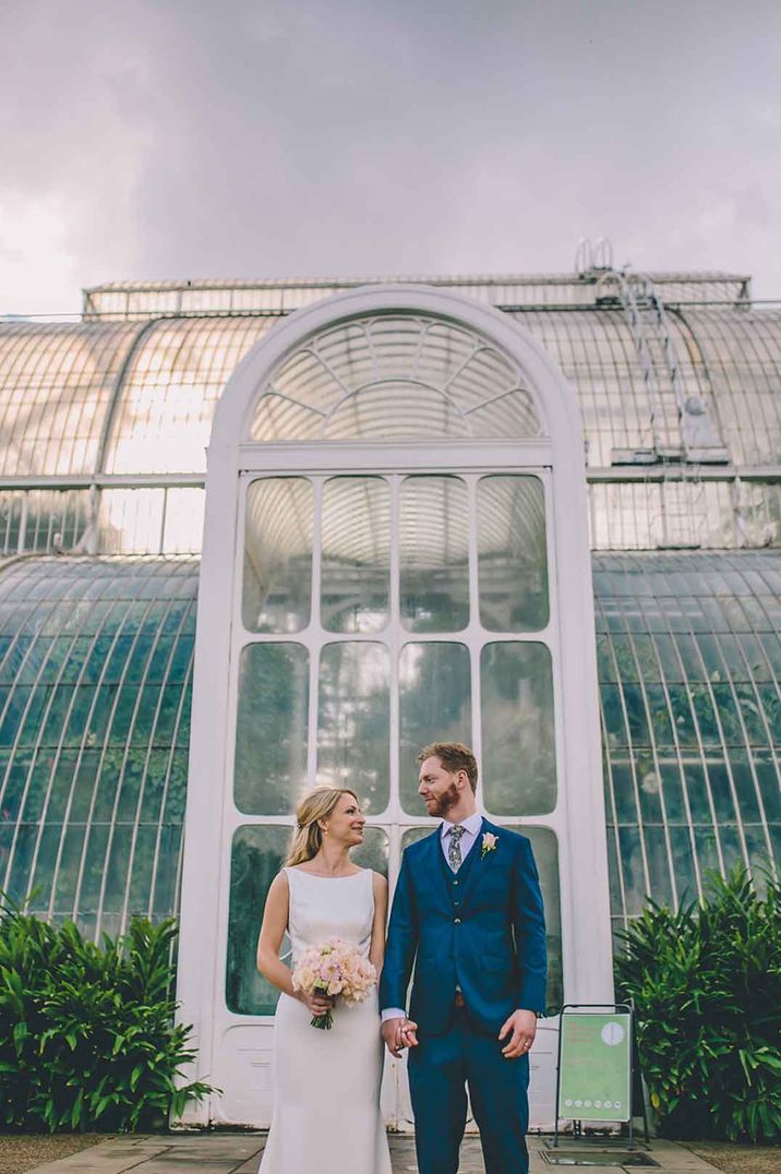 Bride in halter neck sleeveless satin wedding dress standing with groom in blue suit and white boutonniere in front of Kew Gardens London wedding venue 