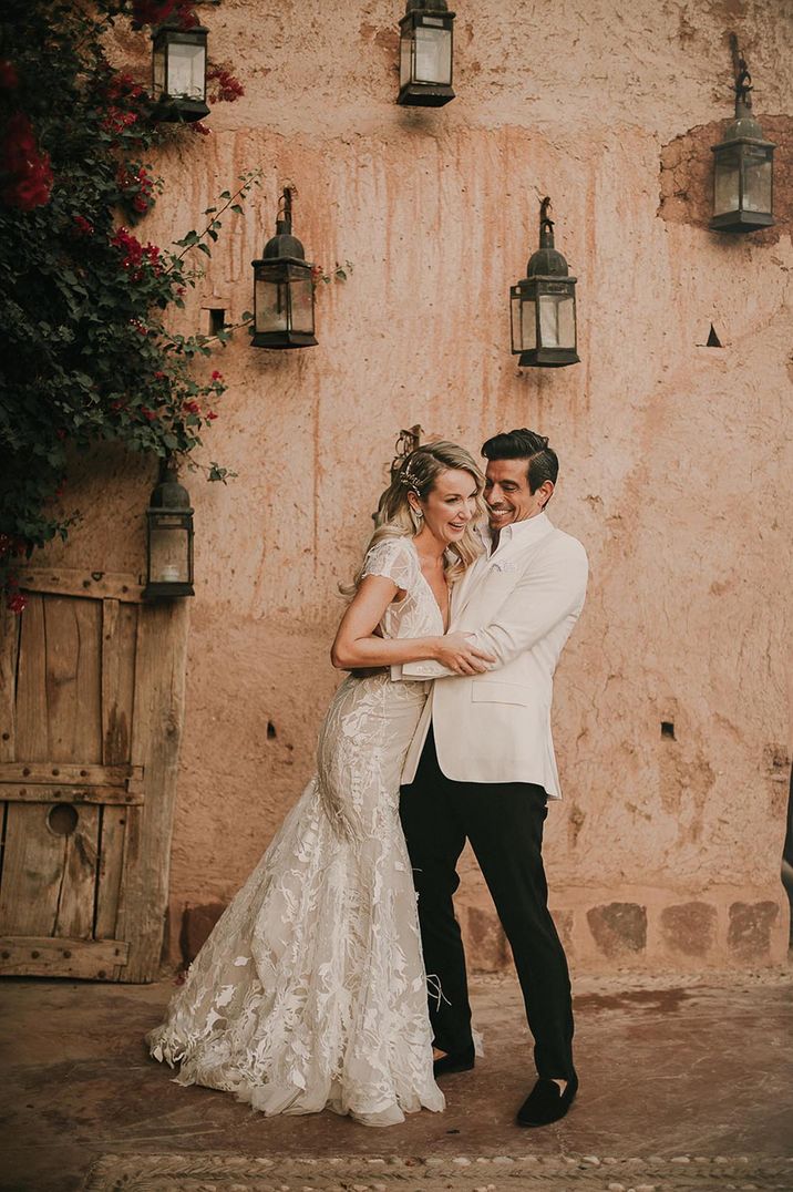 Groom in black trousers with white jacket smiling with the bride in plunging wedding dress with bow detail for wedding over 60k 