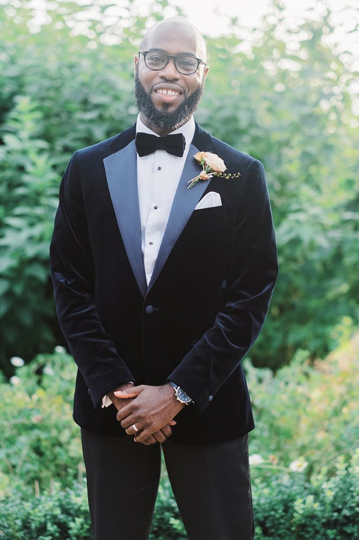 A Black groom with a bald head and full beard stands for wedding portrait wearing black tie.