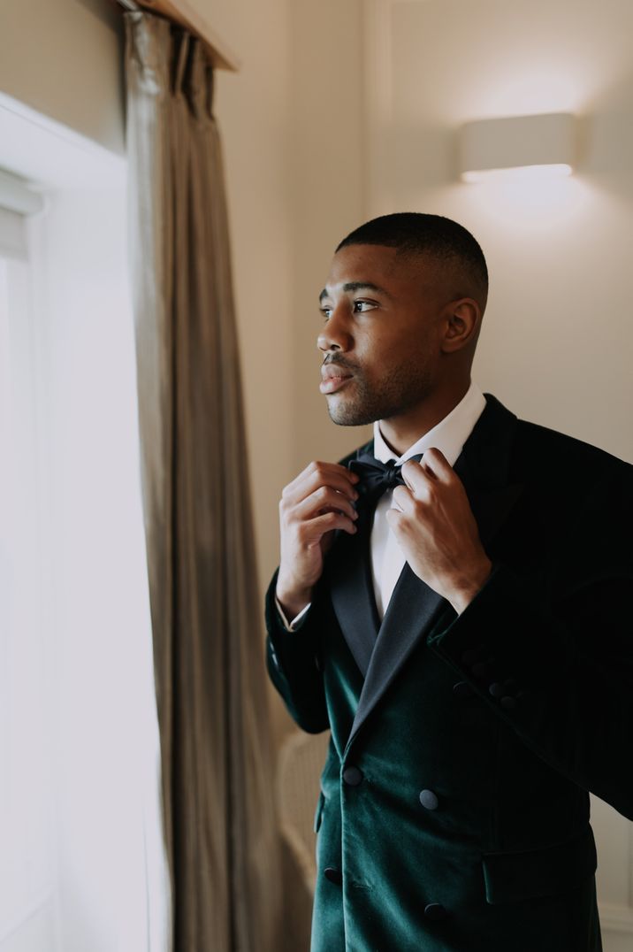 A Black groom wears a dark green velvet tuxedo jacket. He tightens his bow tie.