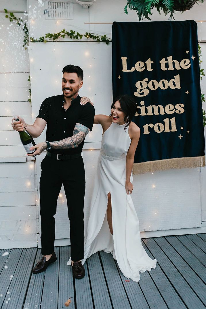 Bride in halter neck wedding dress and groom in black shirt doing champagne pop