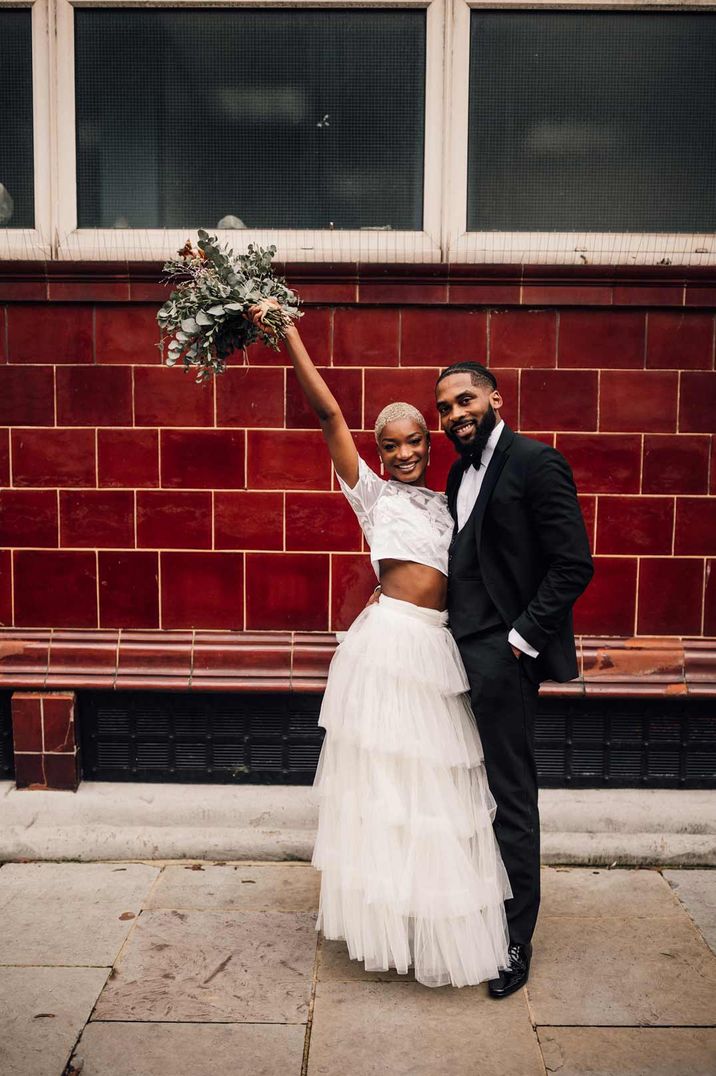 Bride in two piece bridal set with white short sleeve lace crop top and full tulle skirt standing with groom in classic black tuxedo 