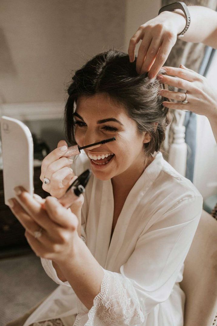 Bride in white lace getting ready robe doing her mascara wedding makeup - timeline of wedding day 