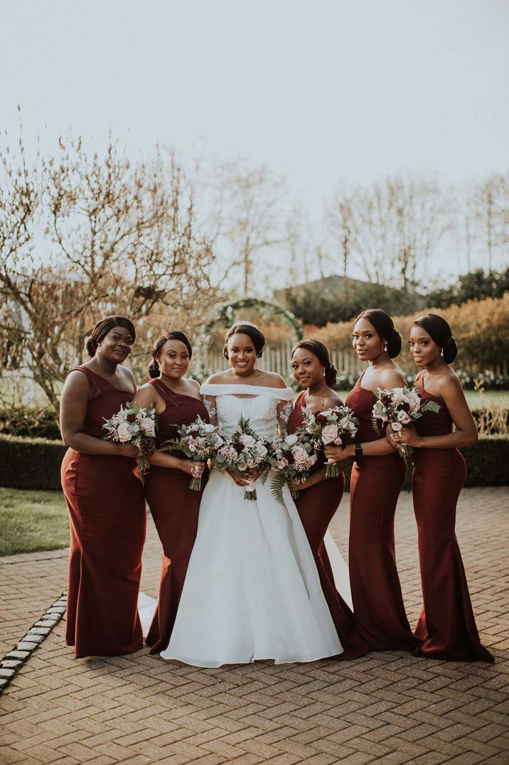 Bride in off the shoulder wedding dress posing with bridal party wearing one strap burgundy bridesmaid dresses 