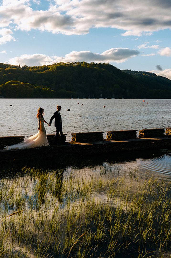 A bride in a mermaid wedding dress and groom walk across the jetty at the Lake District wedding venue 