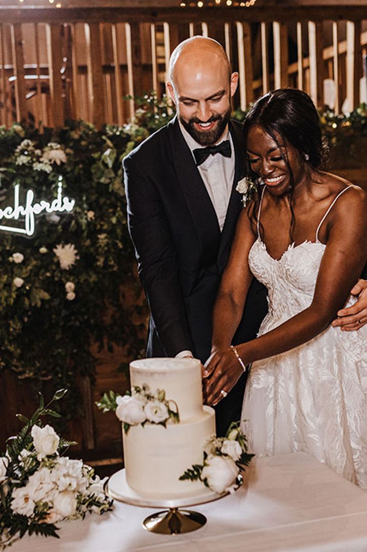 Bride in lace wedding dress and groom in black tuxedo cutting the white wedding cake 