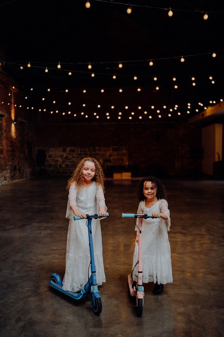 Two girls in white flower girl dresses riding on scooters around the wedding venue 