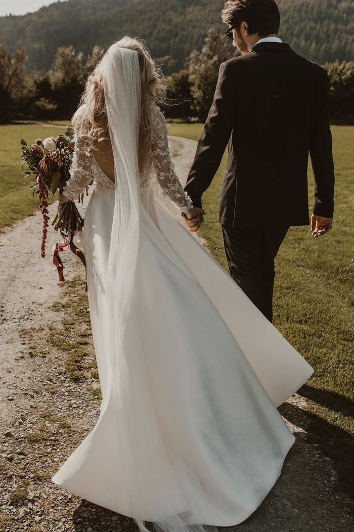 Bride and groom walk away from camera, bride in applique wedding dress with low back and a veil, holding a bouquet, groom in black suit.