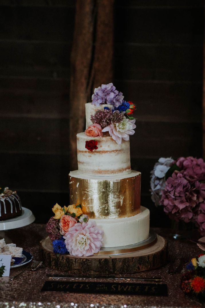 Four tier rustic wedding cake with naked, buttercream and golf leaf layers decorated in colourful flowers 