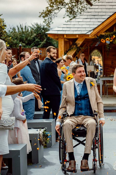 Bride in a lace wedding dress descending up the aisle with her groom in a wheelchair holding a sunflower bouquet 