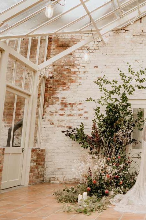 Bride with alopecia wearing rose bridal crown and sparkly strappy wedding dress with sheer bridal wedding wings and groom in black tuxedo standing amidst the foliage, fauna and dried flowers at Our Beautiful Glasshouse venue 