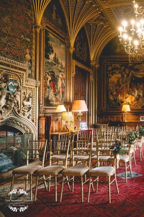 Eastnor Castle gothic drawing room with dark red carpet, large golden chandelier, large golden framed historic portraits and foliage aisle decor 