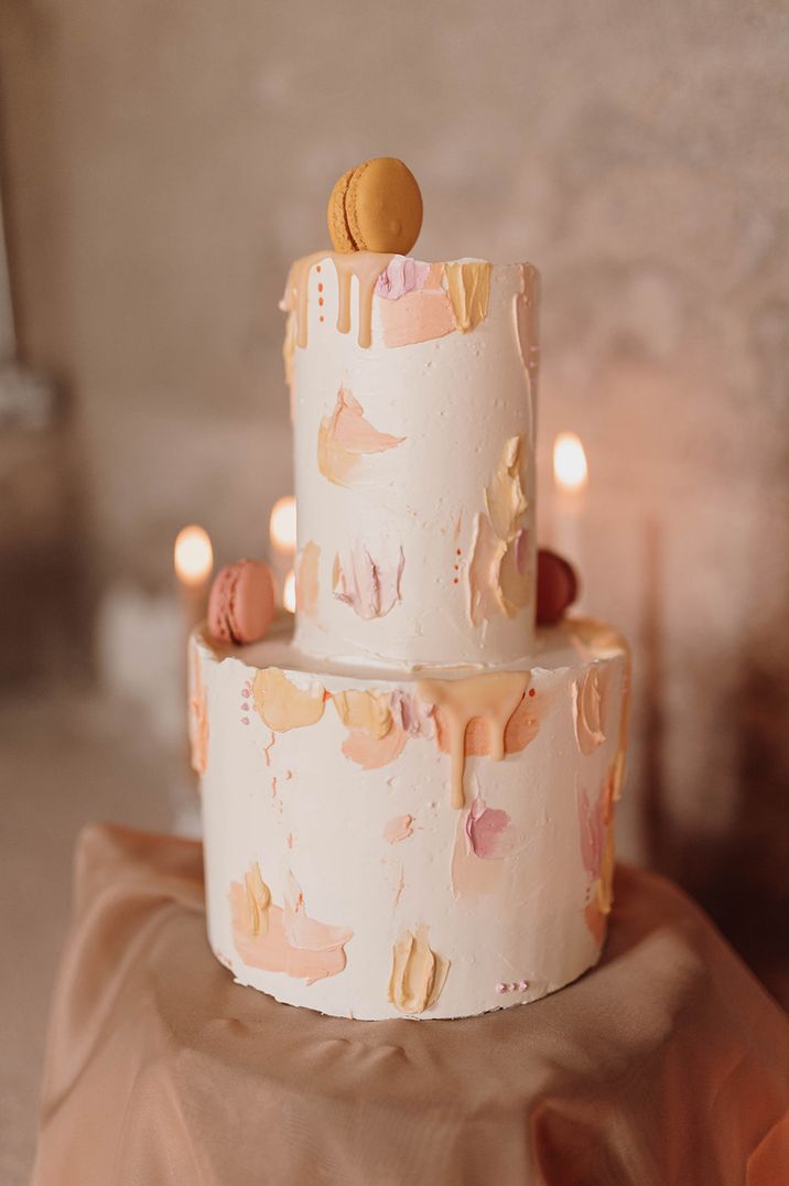 Two tier white iced wedding cake with pastel pink, yellow and orange painted pattern with drip detail and macarons