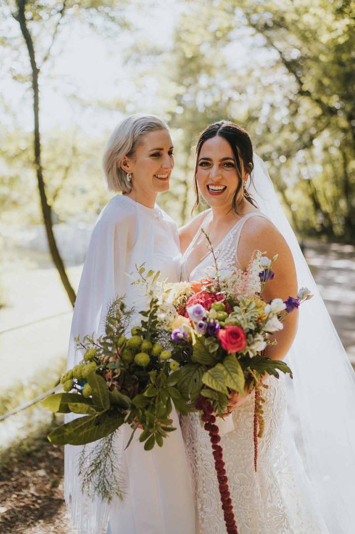 Bride in v neck wedding dress and church length veil standing with bride in bridal jumpsuit with cape both holding large wildflower and colourful floral bouquets - wedding budget spreadsheet 