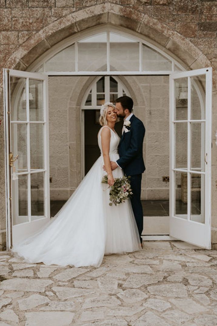 Bride in boatneck sleeveless wedding dress holding dried flower bridal bouquet being embraced by groom in classic black tuxedo at Pennsylvania Castle Estate wedding venue 