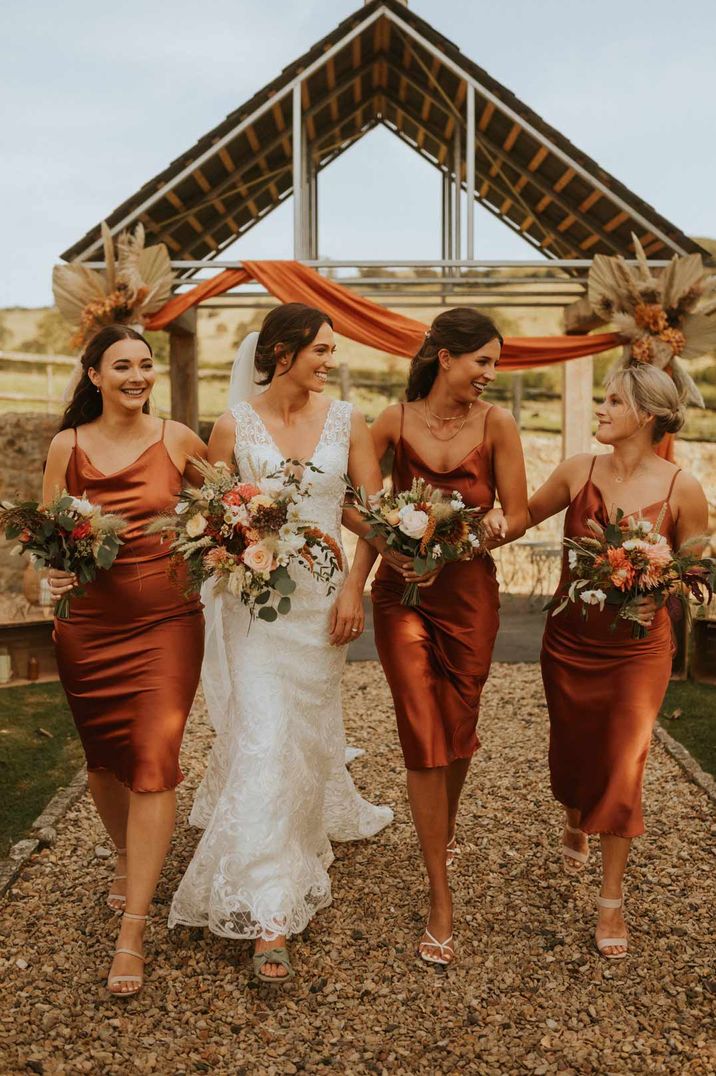 Bridesmaids in burnt orange satin midi cowl neck bridesmaid dresses walking with bride in lace wedding dress 