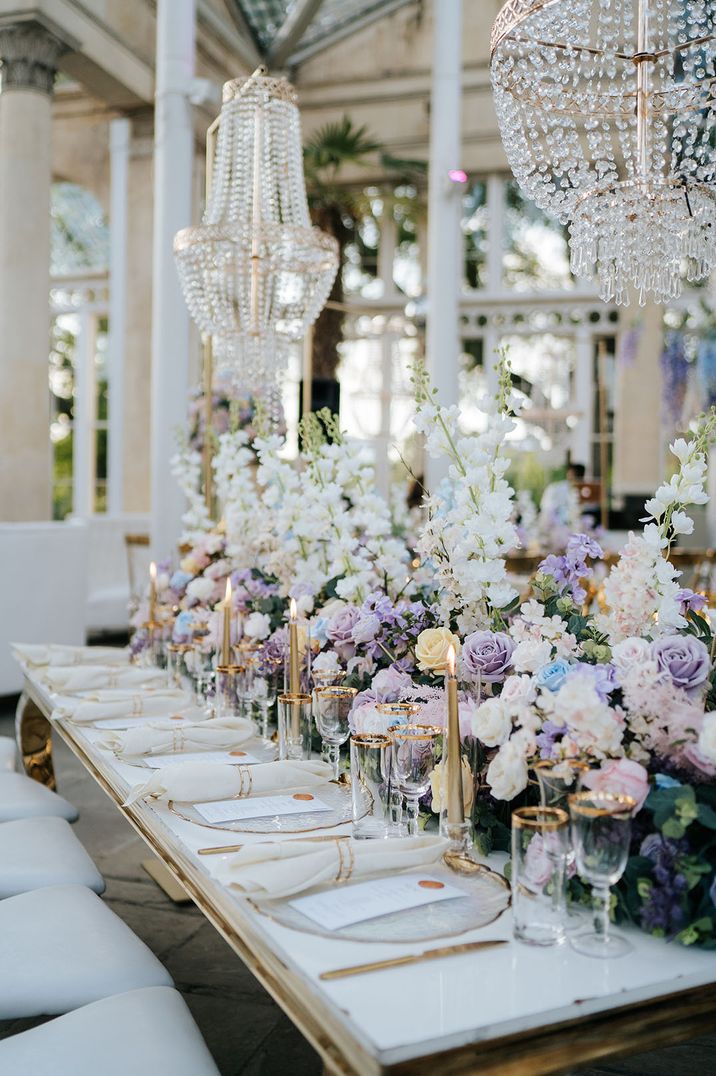 Lilac, white and light blue pastel wedding flowers decorating the wedding tablescape with gold candles and chandeliers 