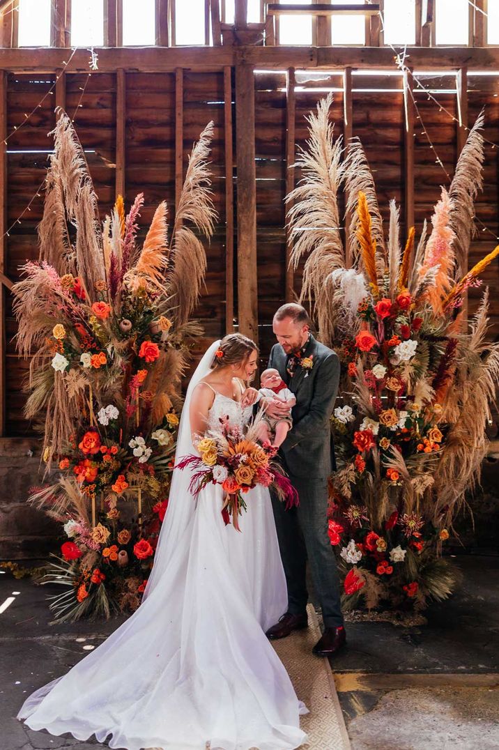 Bride in floral lace strappy wedding dress, groom in grey suit and boutonniere holding their baby amidst vibrant floral arrangement