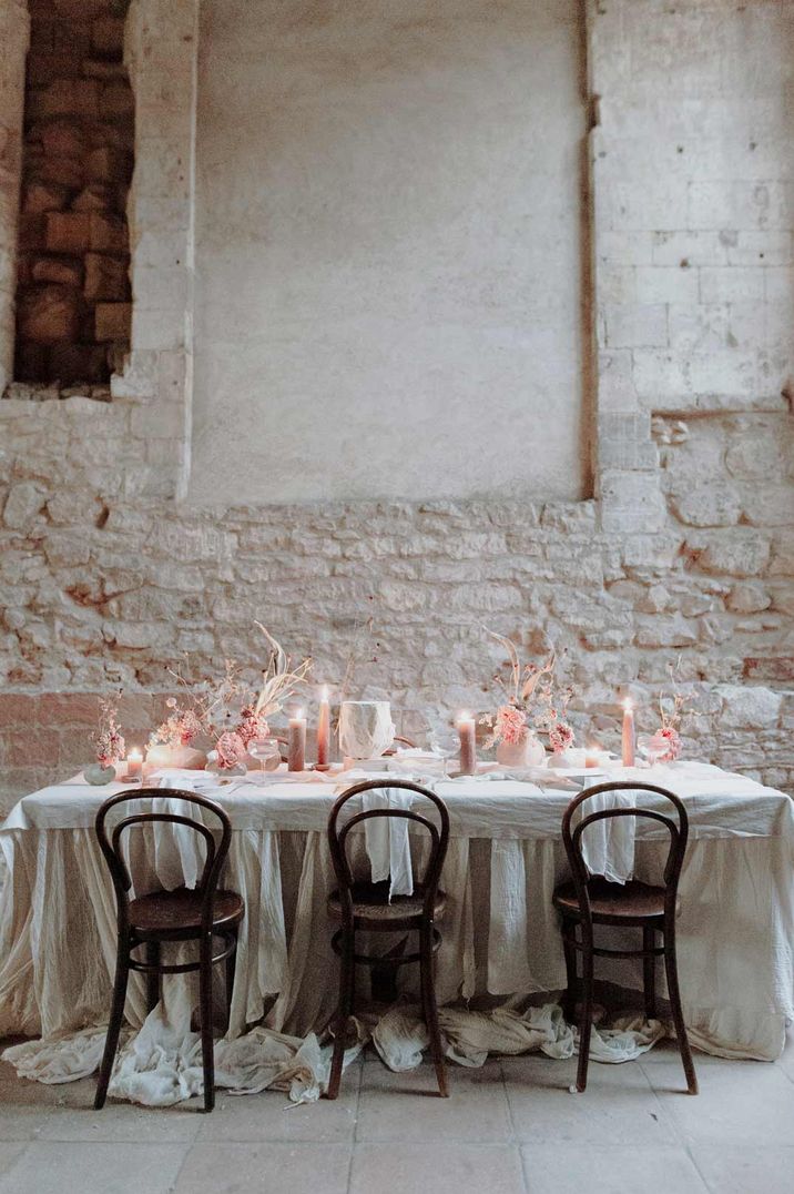 Neutral and rustic wedding tablescape with white cotton tablerunner, dark mahogany chairs, neutral pink tapered and pillar candles and pink dried floral decorations with exposed brick at Blackfriars Priory