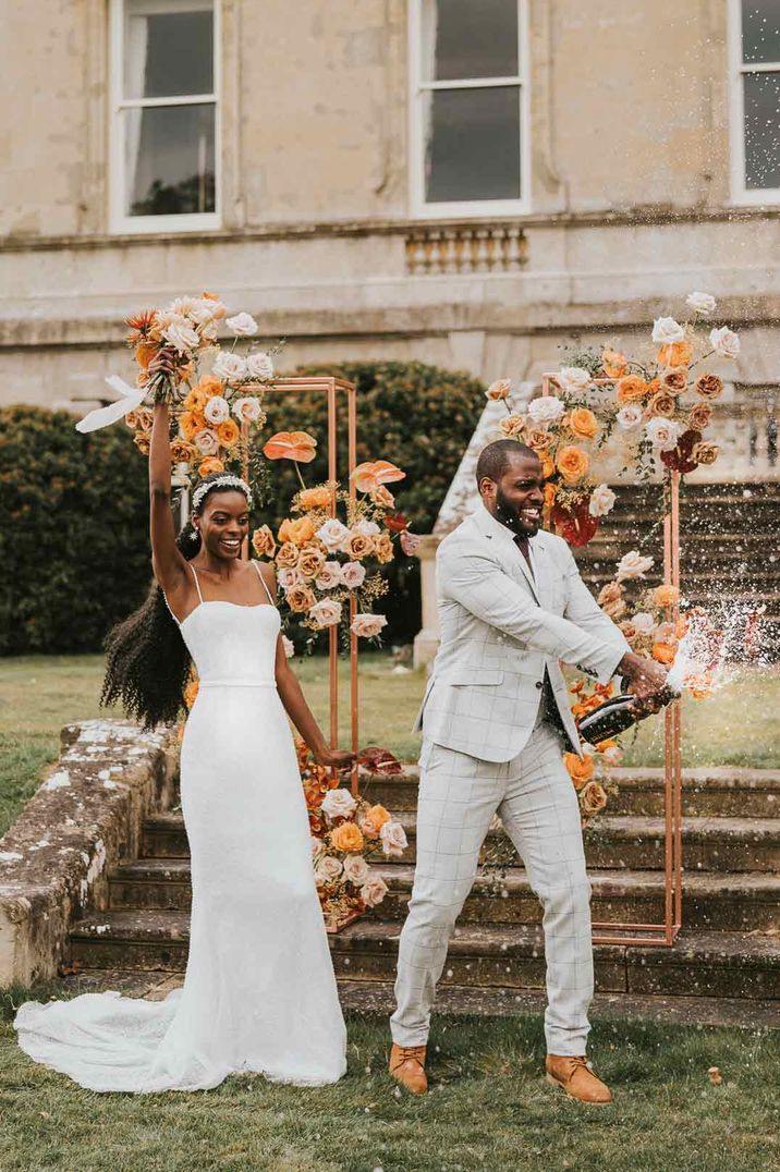 Bride in strappy satin wedding dress and groom in light grey suit doing champagne pop wedding photo at Kirtlington Park wedding venue 