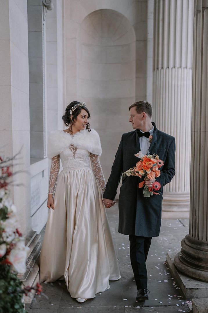 The bride in a long sleeve lace wedding dress with small princess skirt walks with the groom in traditional black tuxedo