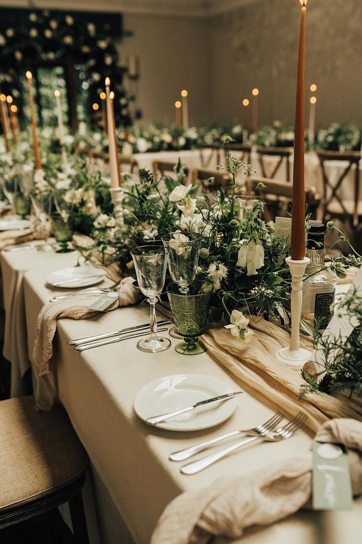 Cream tablecloth and runner with lots of foliage and brown and cream taper candles with green glassware for botanical style wedding 