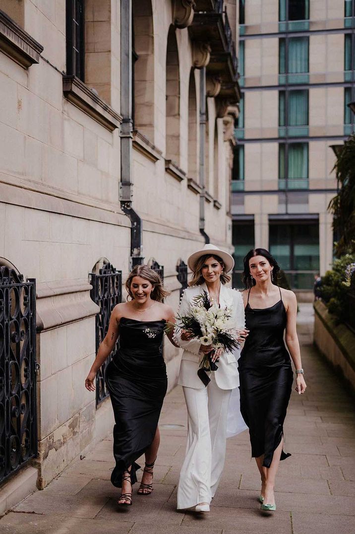 Bride in white bridal suit with fedora hat walking with bridesmaids in strappy black bridesmaid dresses 