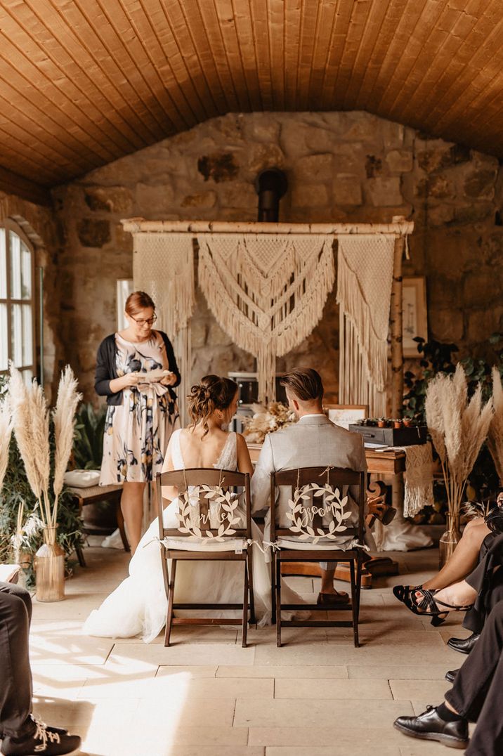 Rustic wedding ceremony in an orangery with macrame backdrop, pampas grass in cases and wooden chair back signs 