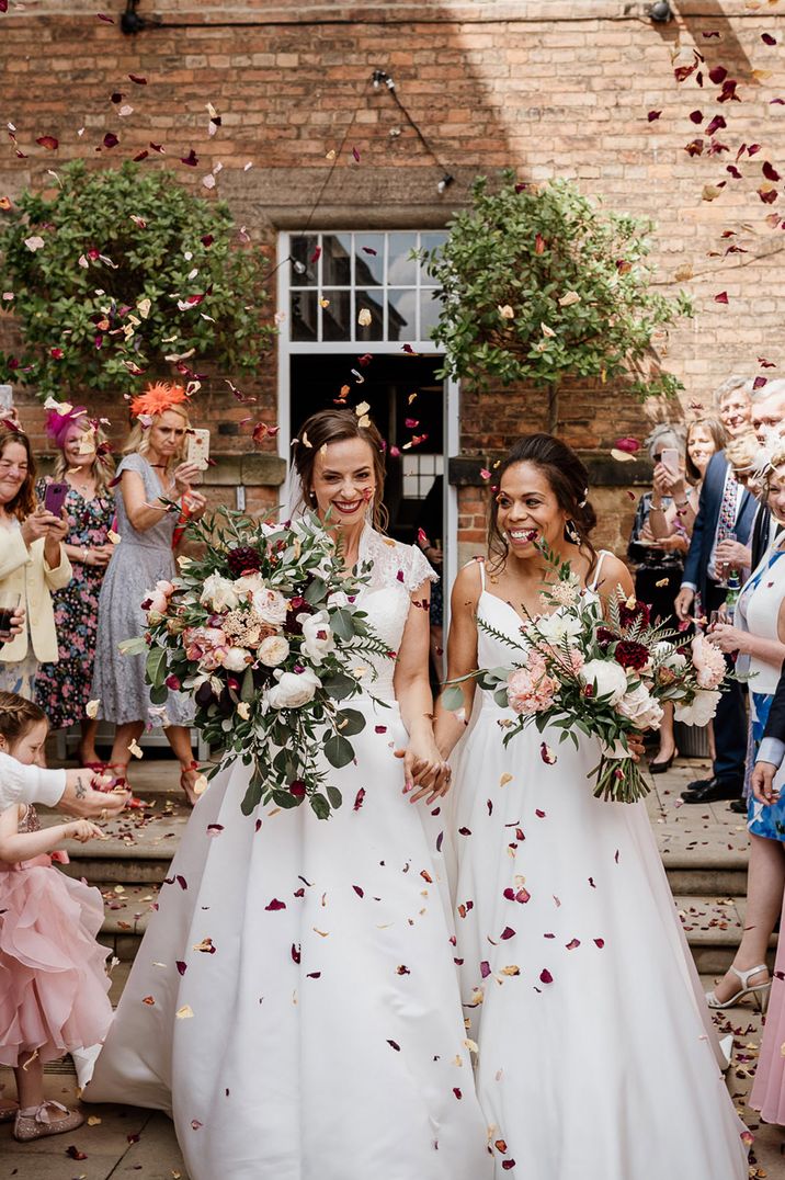 Confetti moment with brides in lace and satin dresses holding giant bouquets at West Mill Derby 