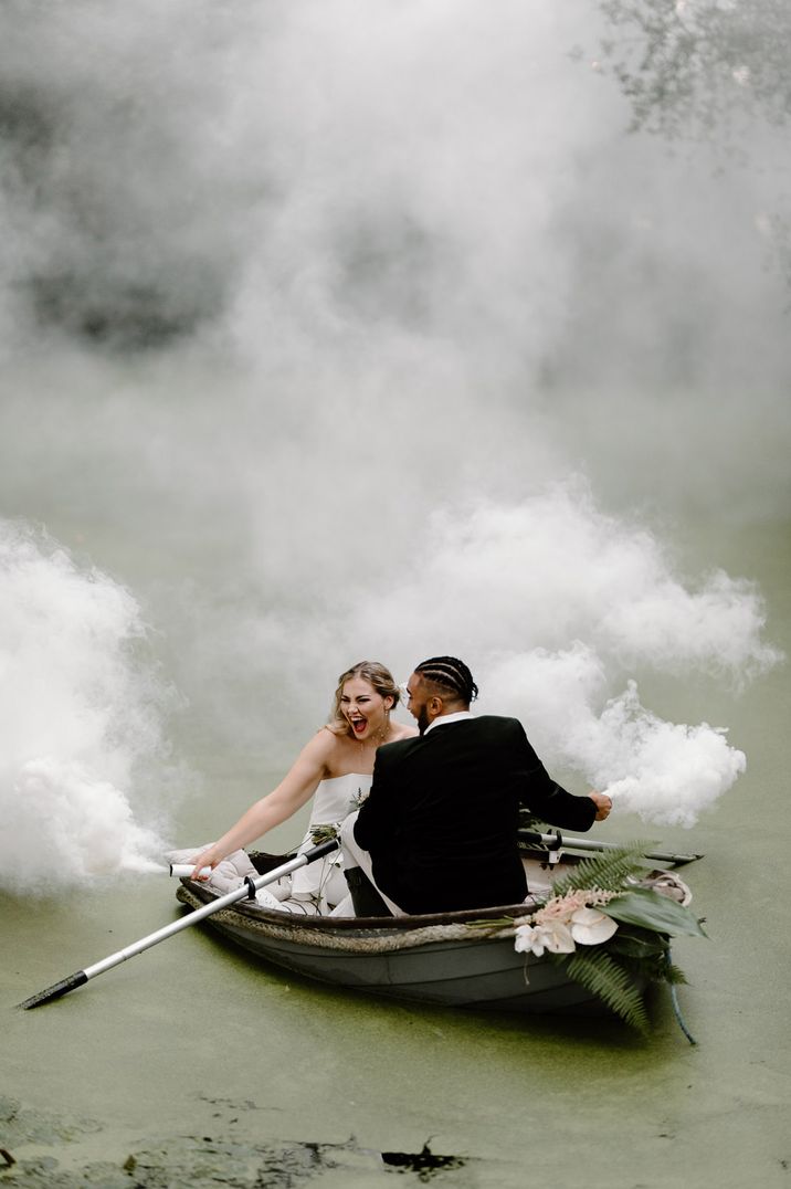 Bride and groom sitting in a boat with white smoke bombs going off around them 