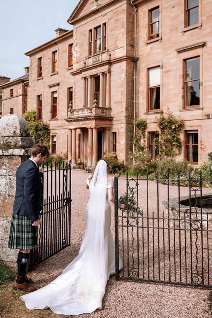 Bride in off shoulder satin wedding dress with church length bridal veil walking through the gate with groom in wedding kilt at Cambo House wedding venue Scotland 