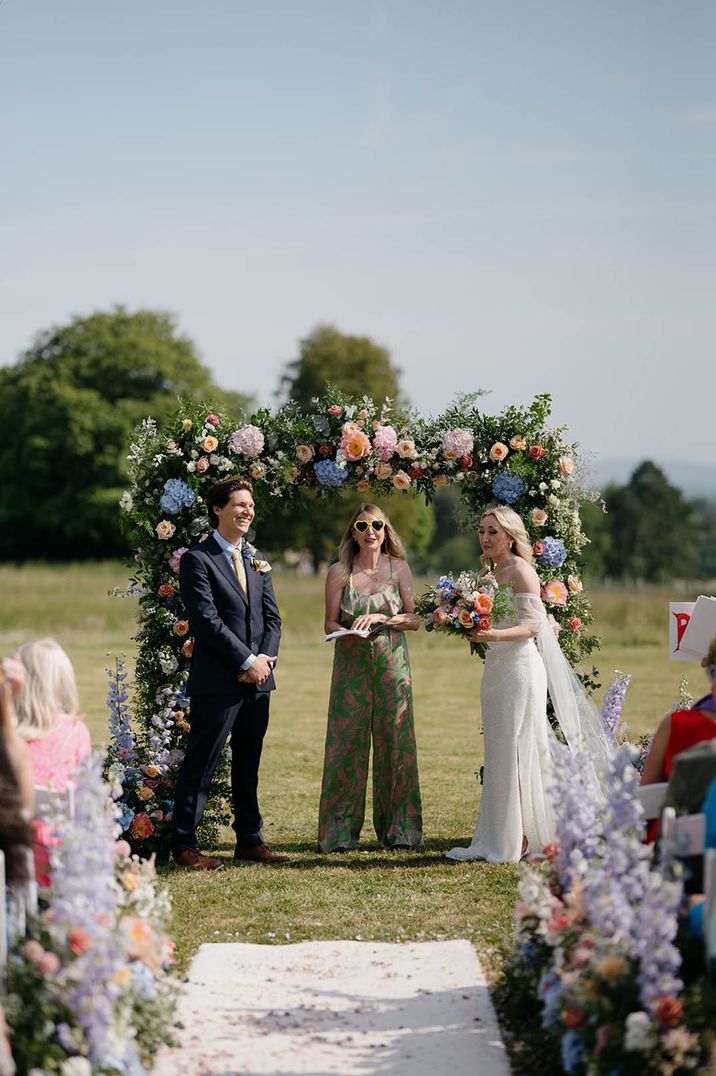 Purple, orange and blue wedding arch altar flowers for outdoor wedding with wedding celebrant in sunglasses 