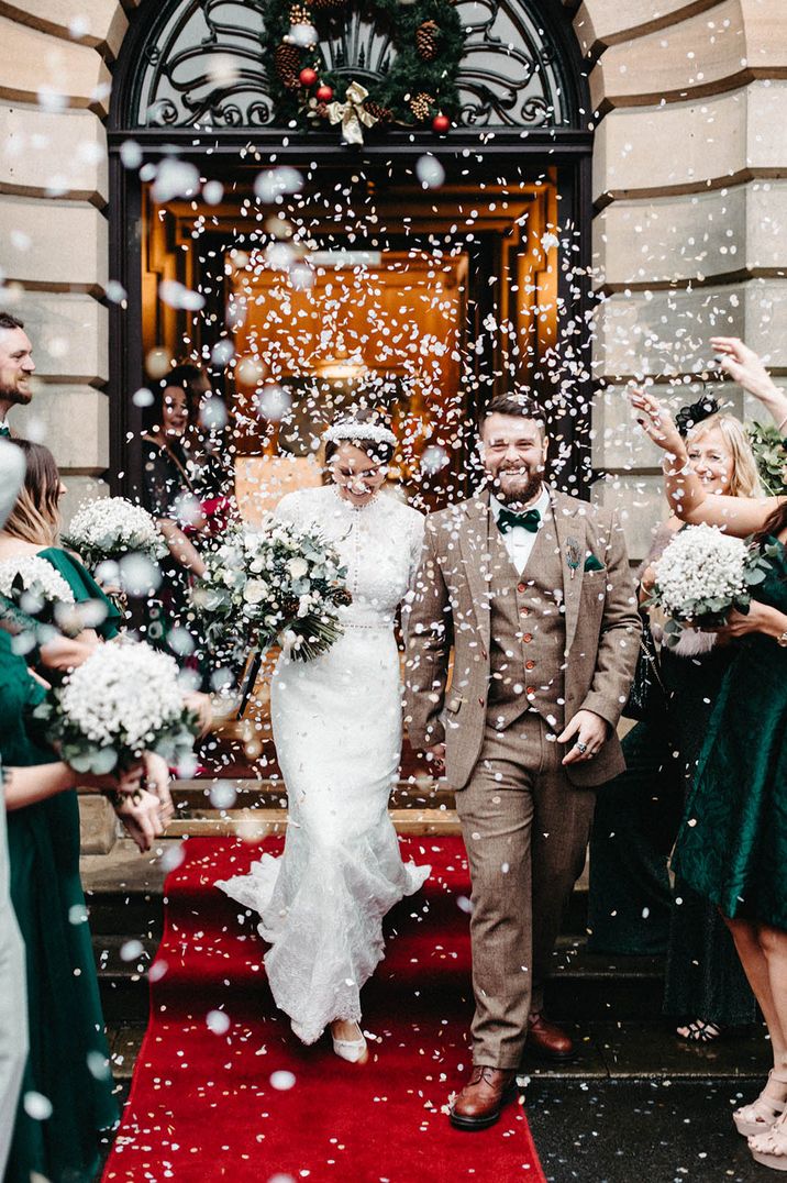 Groom in tweed suit walking with the bride in a lace long sleeve wedding dress for their confetti exit 