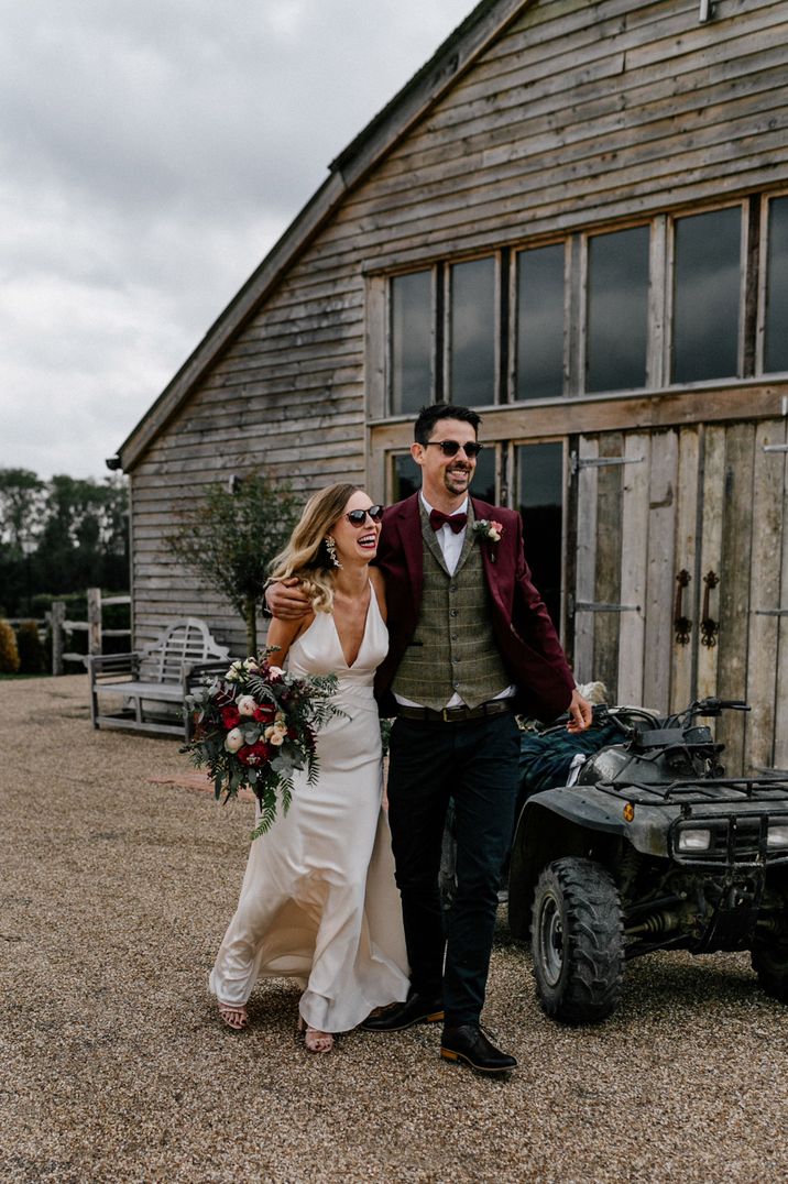 Groom in a burgundy suit, bow tie and patterned waist coat with bride in satin wedding dress with a quad bike 