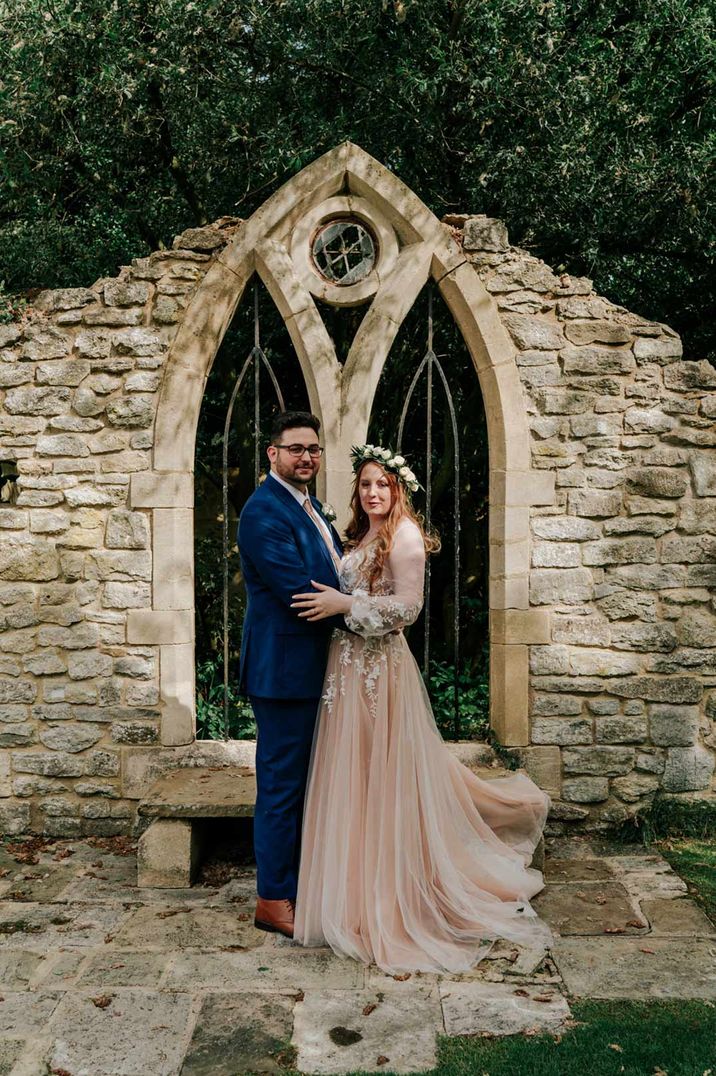 Bride in pink long sleeve lace detailed wedding dress with white rose flower crown and groom in dark suit embracing