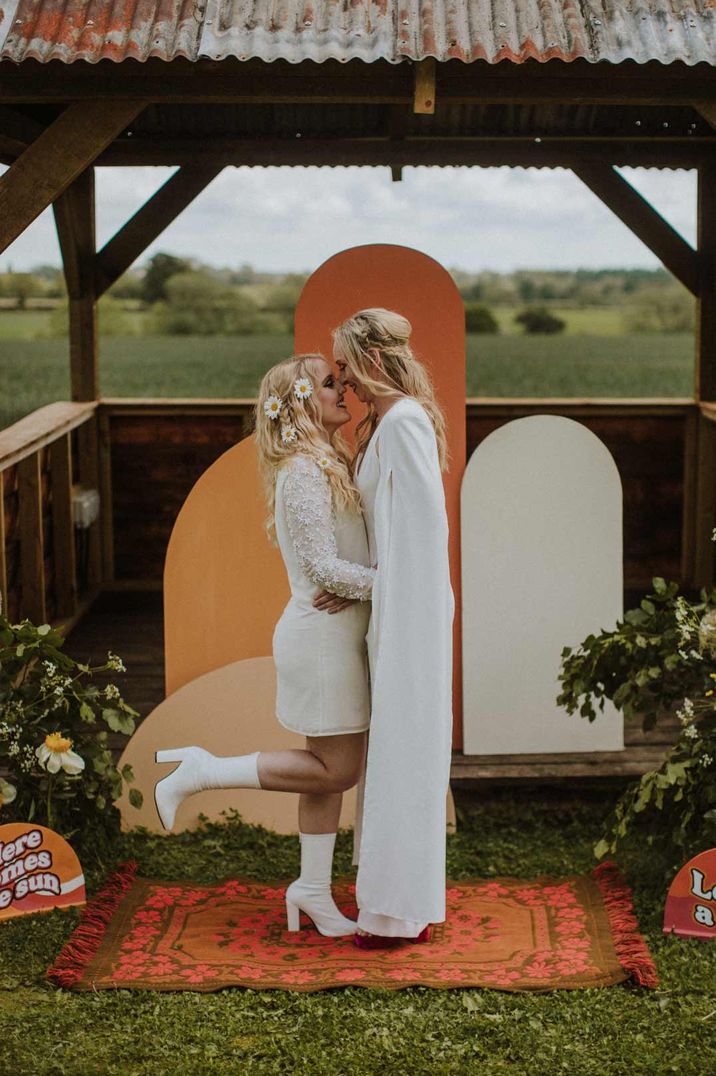 Bride in short long sleeve lace wedding dress and large daisy floral hair accessories with white ankle boot heels and bride in boho long sleeve wedding dress embracing in front of burnt orange, off-white and mustard rounded wedding backdrop