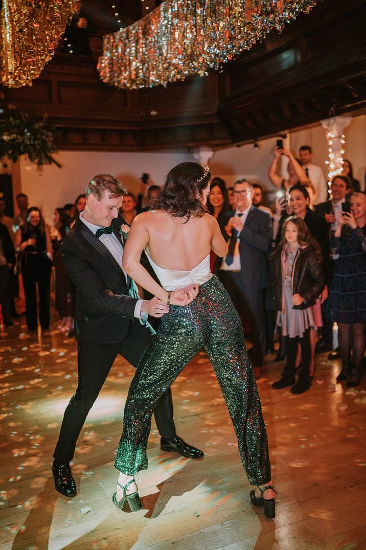 The bride in a low back crop top and dark green sequin trousers with matching green block heels dances on the dancefloor with the groom 