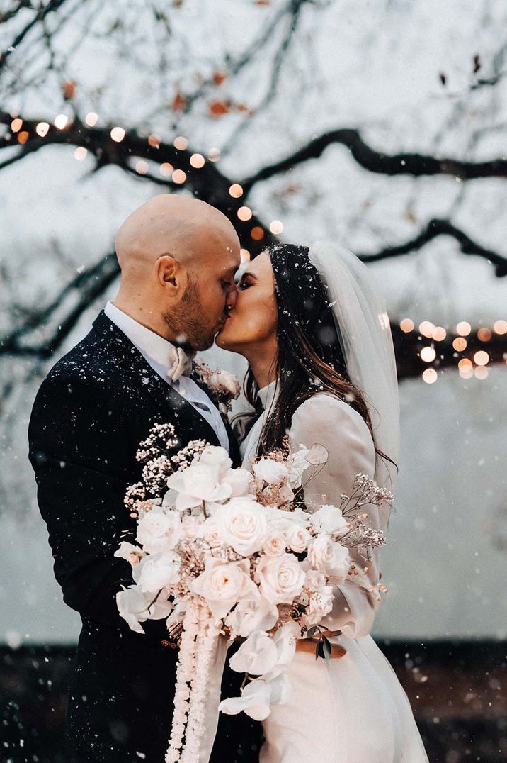 Bride wearing long sleeve wedding dress carrying white flower wedding bouquet kissing the groom as snow falls around them 