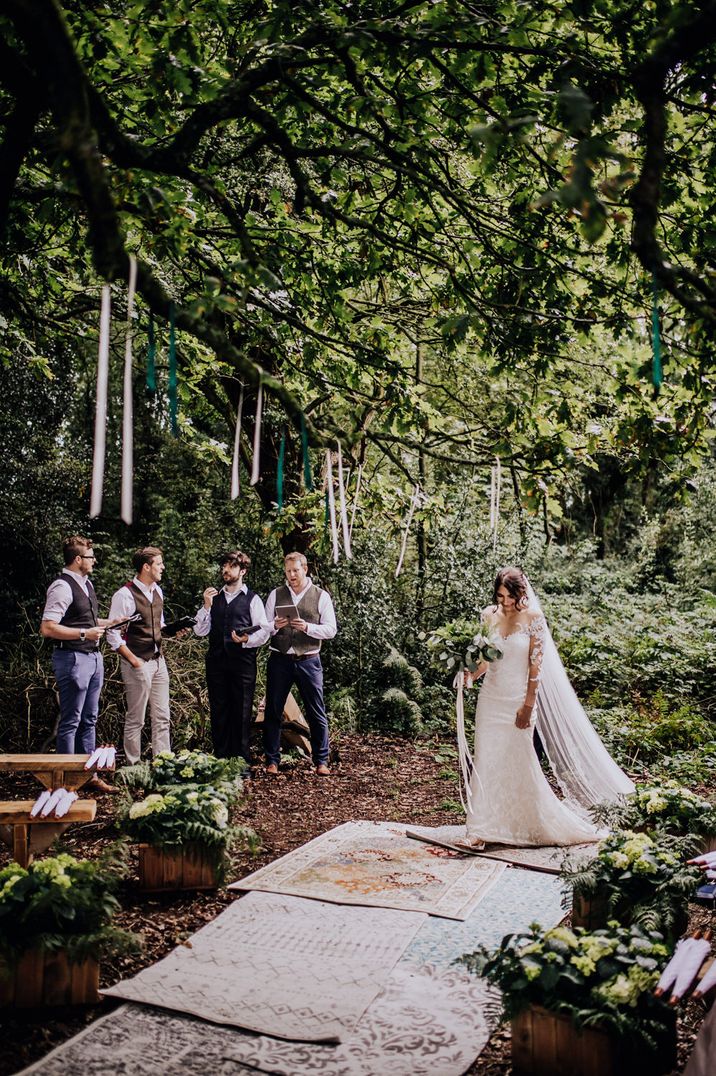 Bride in a long sleeve lace wedding dress walking down the wooden wedding aisle on Persian rugs with streamers in the trees and the groomsmen in waistcoats 