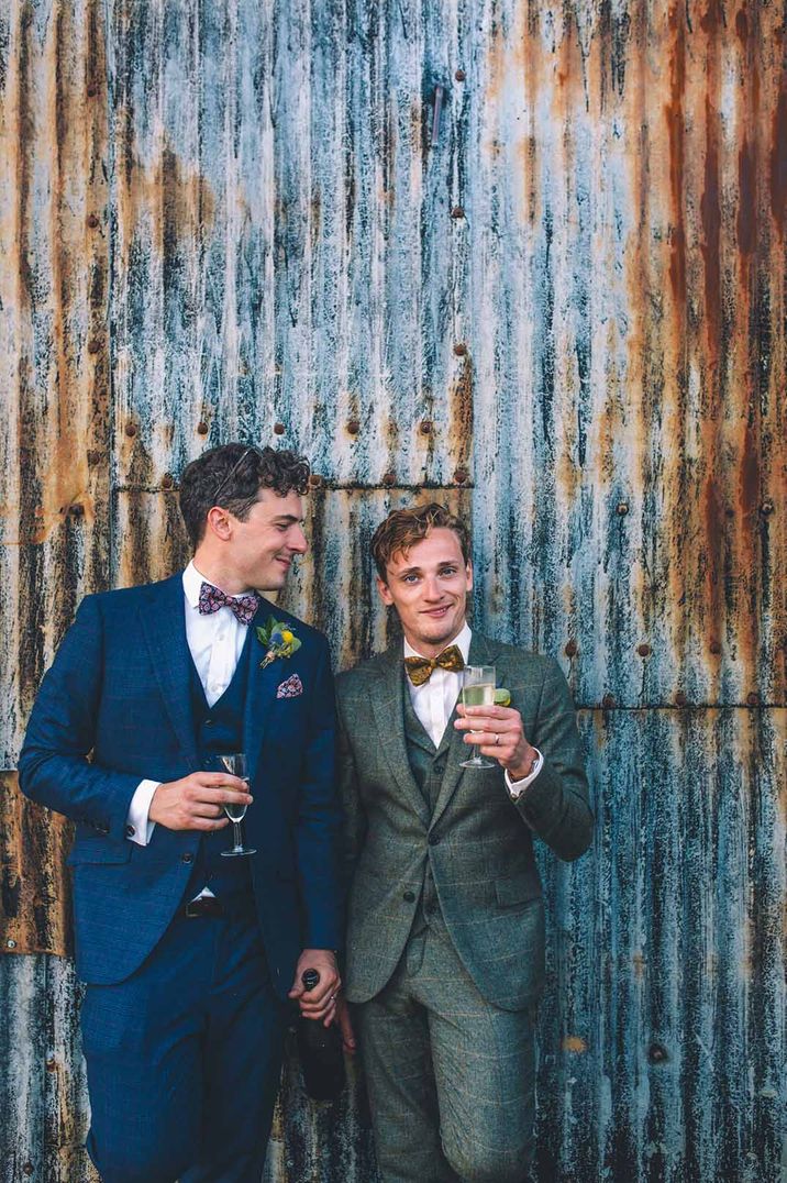 Grooms stand beside industrial style wall with drinks and laughter 