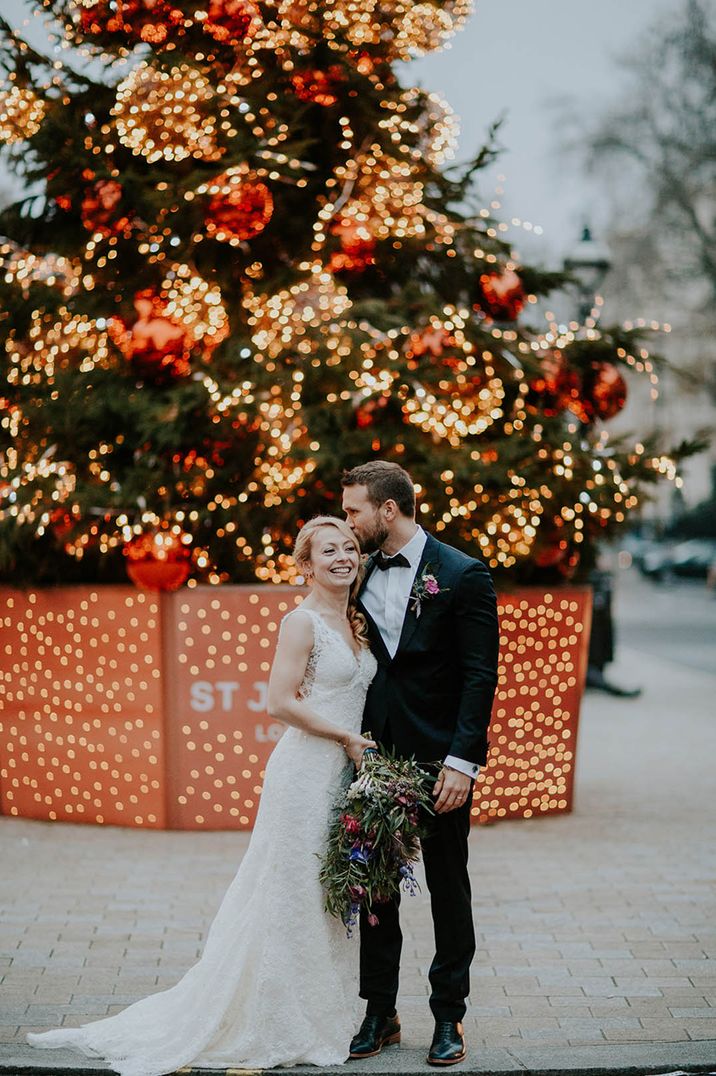 Groom in black tie kisses the bride in a lace wedding dress on the forehead in front of large Christmas tree in London 