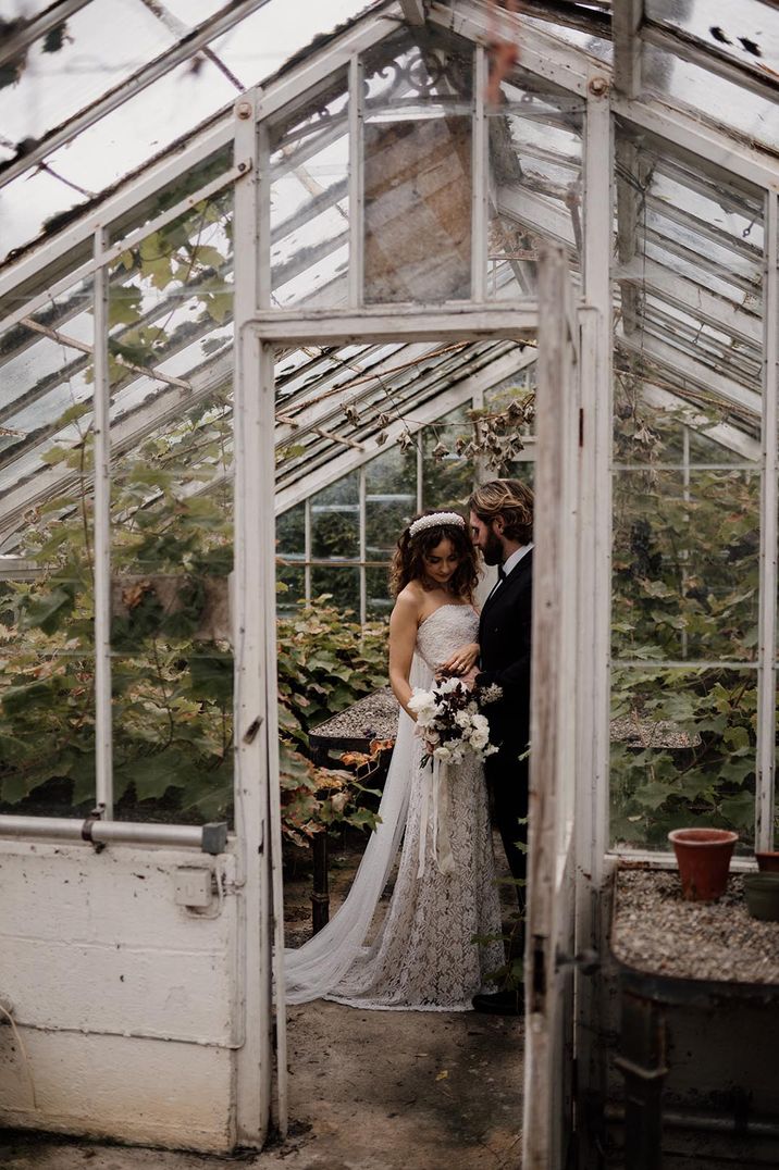 Bride in strapless lace wedding dress and groom in dark tuxedo standing in glasshouse wedding venue 