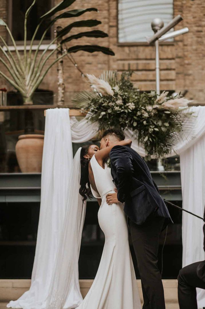 Bride in sleeveless satin wedding dress kissing groom in black tux at alter of Devonshire Terrace London wedding venue 