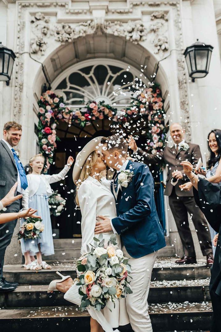 Bride in long sleeve white satin suit set and bridal hat kissing groom in navy blue suit at small intimate wedding 