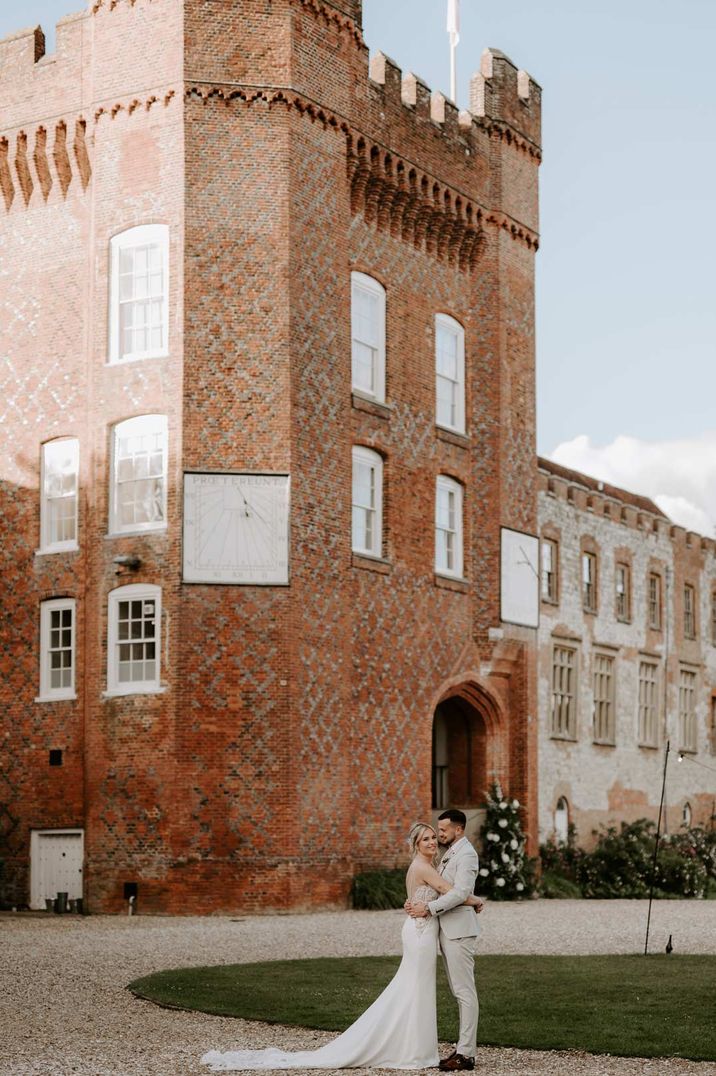 Bride in strapless wedding dress and groom in light grey suit embracing outside of Farnham Castle wedding venue 