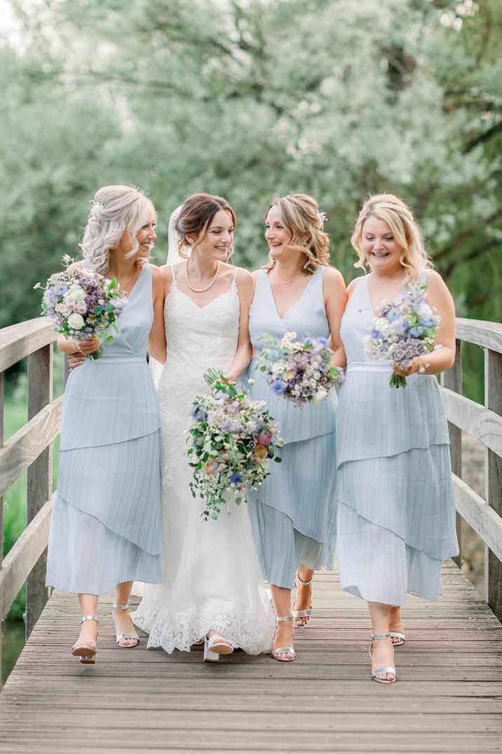 Bride in strappy white wedding dress walking over bridge with bridesmaids wearing sleeveless layered blue bridesmaid dresses 