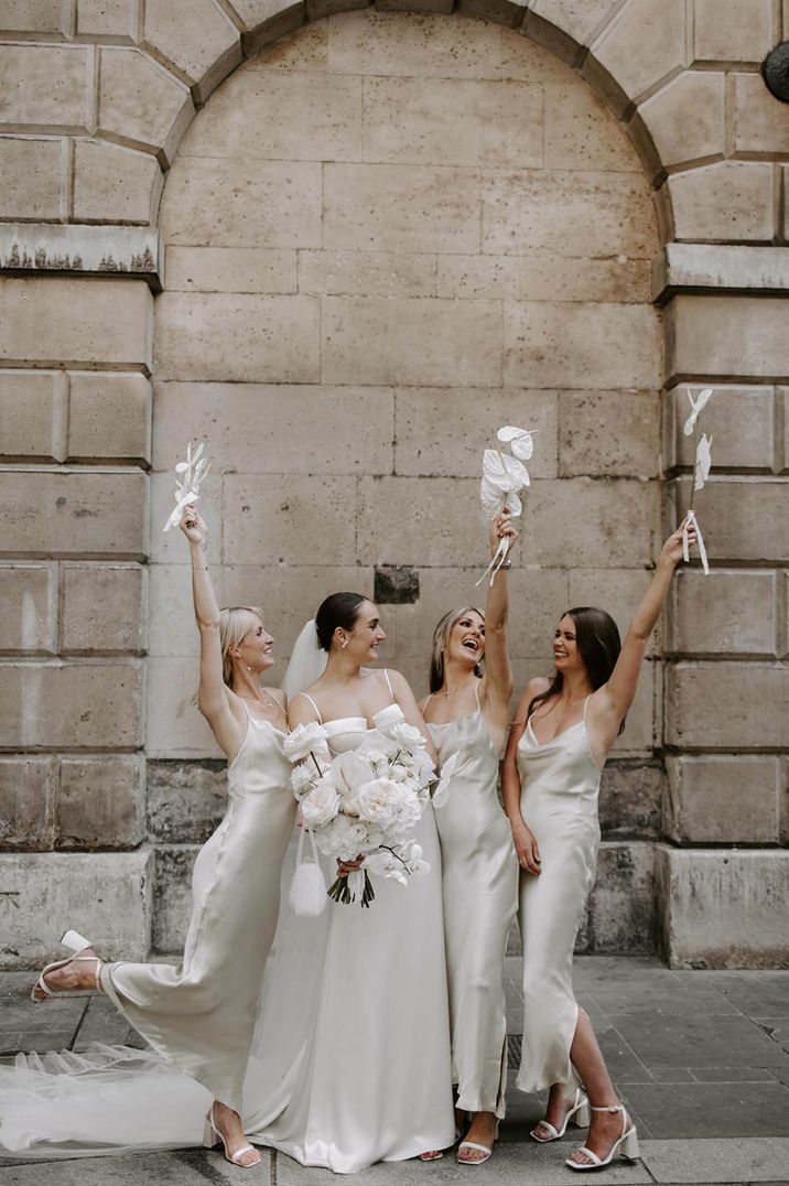 Bride in off the shoulder satin wedding dress standing with bridal party in sleeveless satin white bridesmaid dresses 