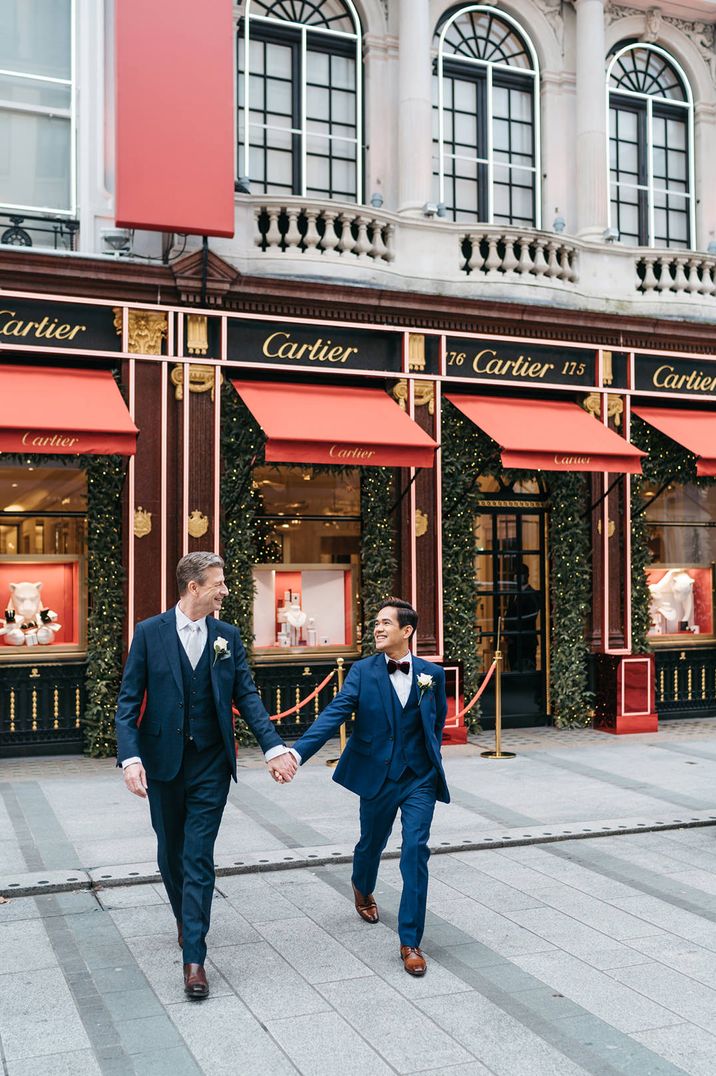 Two grooms in navy three-piece suits holding hands outside Cartier at their London Elopement 