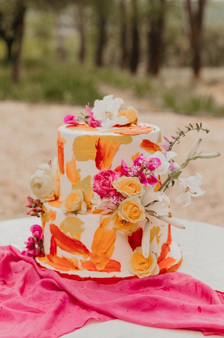 Two tier white iced wedding cake with orange, red and yellow painted pattern with pink and white flower decoration 